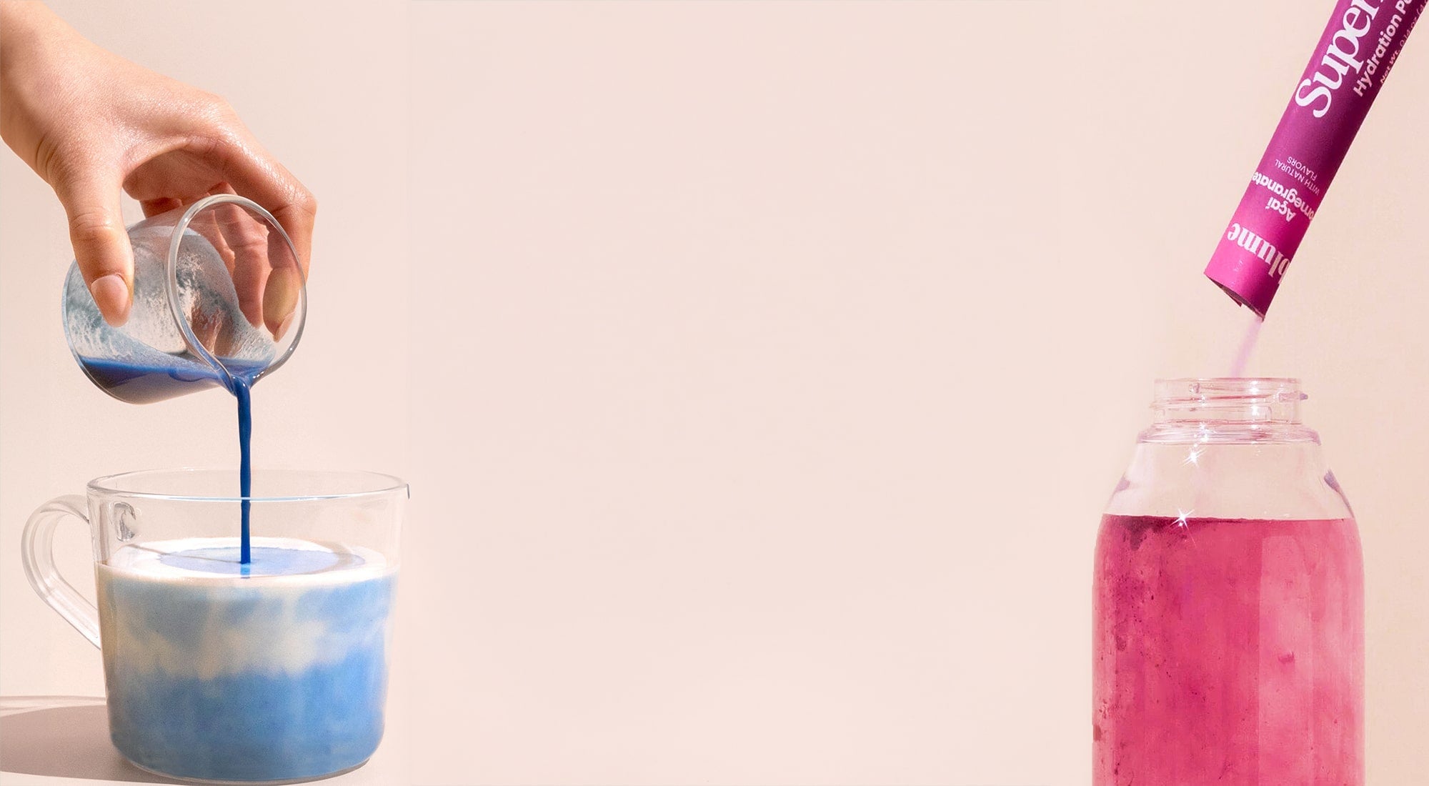 A hand pours blue liquid into a glass mug with frothy white liquid inside. To the right, a pink drink mix stick is being added to a jar filled with pink liquid against a beige background.